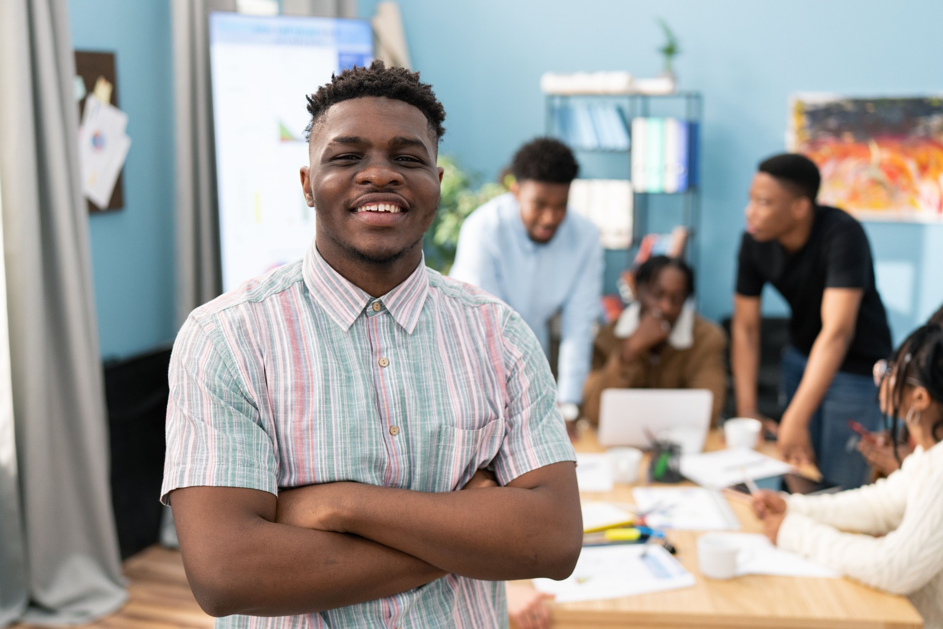 Portrait of a dark-skinned man wearing collared shirt, he smiles showing white teeth, he has black short-cropped hair, he crosses his arms on chest, in the background corporate friends form reports